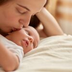 Mother and her newborn baby together lying on bed on blanket. Happy mother kissing and hugging baby, kid looking away and studying outward things. Maternity and parenthood.