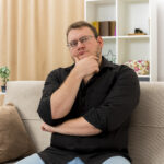 thoughtful adult slavic man in optical glasses sits on armchair holding chin and looking at side inside designed living room